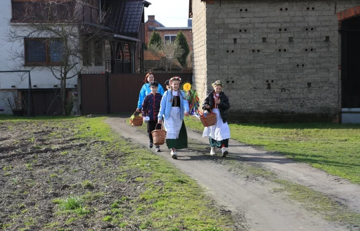 Czworo dzieci w strojach regionalnych, trzymają nowe lotko i koszyczki na dary. Droga gruntowa, w tle zabudowania.
